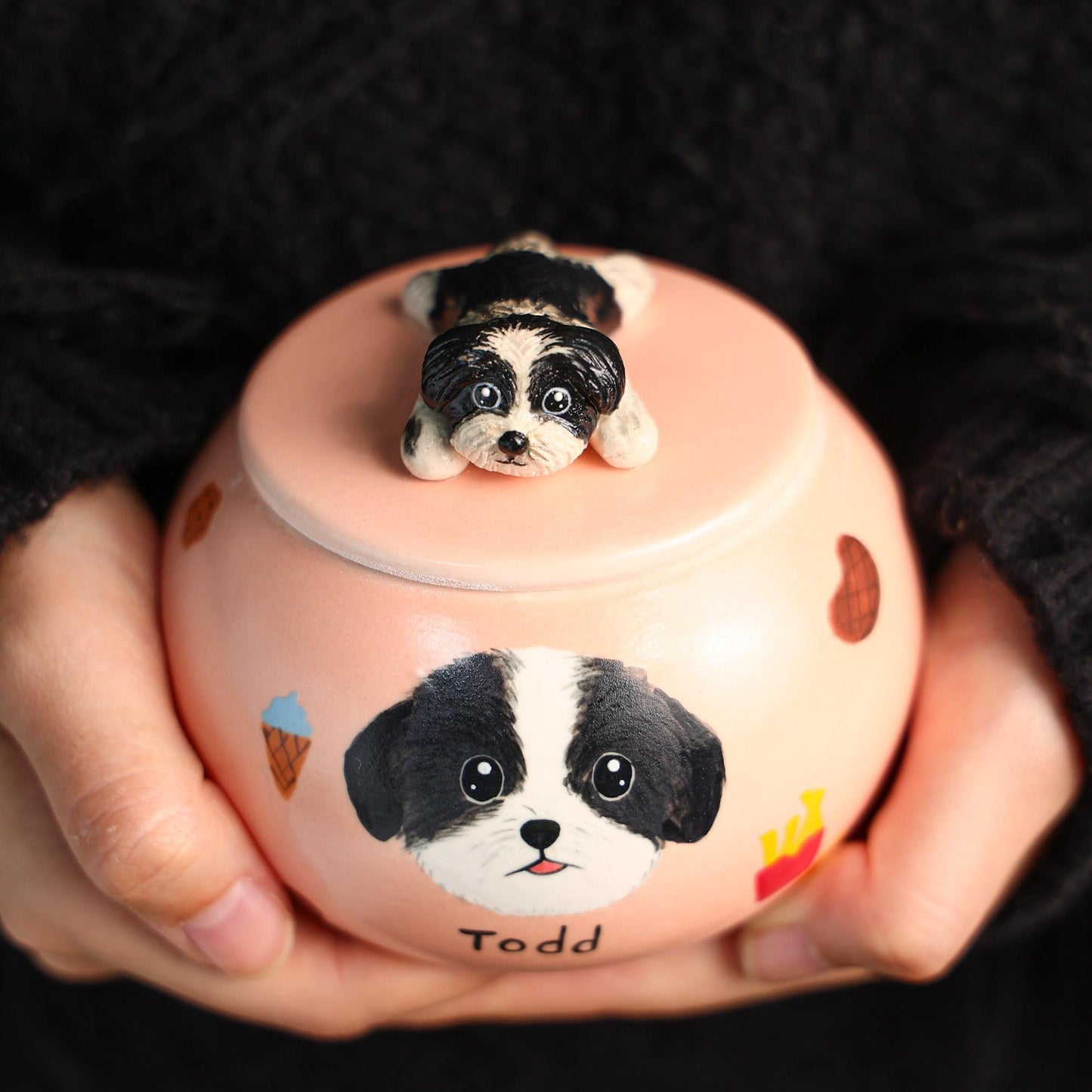 Pink ceramic urn with a dog design and a small dog figurine on top, held by hands against a dark background.