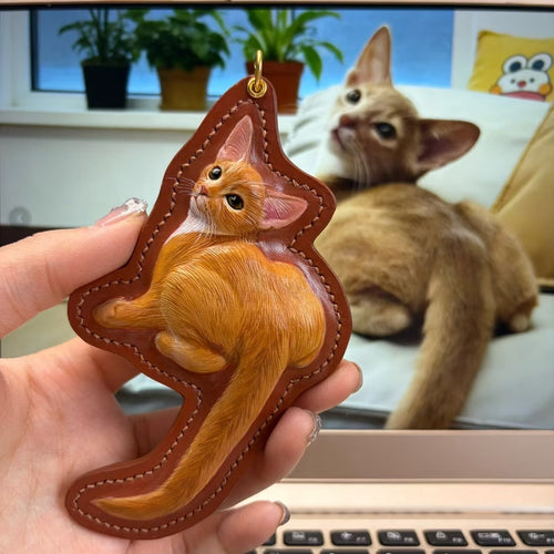 Hand holding a custom pet leather cat-shaped keychain with a cat in the background