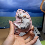 Hand holding a pink Leather Pet Portrait Keychain with a cat design against a computer screen background.