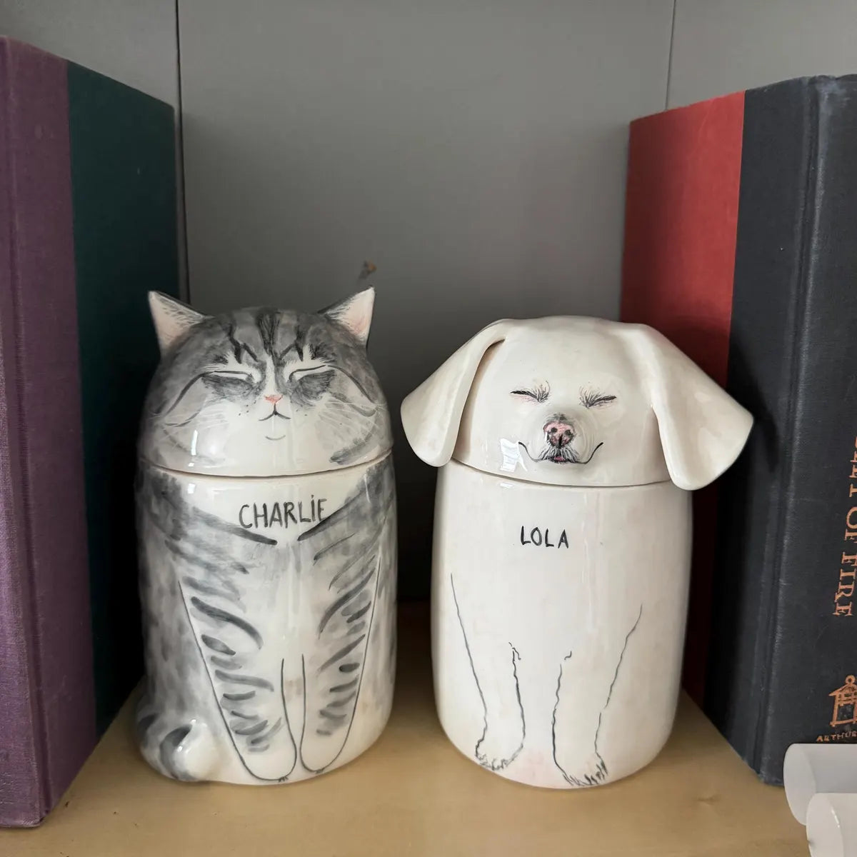 Two ceramic jars shaped like a cat and a dog with names 'Charlie' and 'Lola' on a shelf.