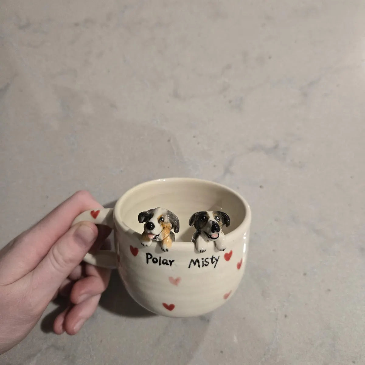 Ceramic mug with two dog figures and 'Polar Misty' text, held by a hand on a neutral background