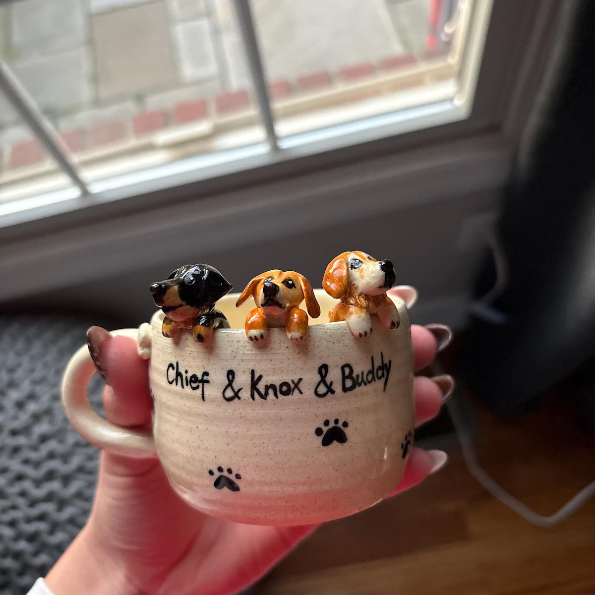 Ceramic mug with three dog figurines and names 'Chief', 'Knox', and 'Buddy' on a blurred background.