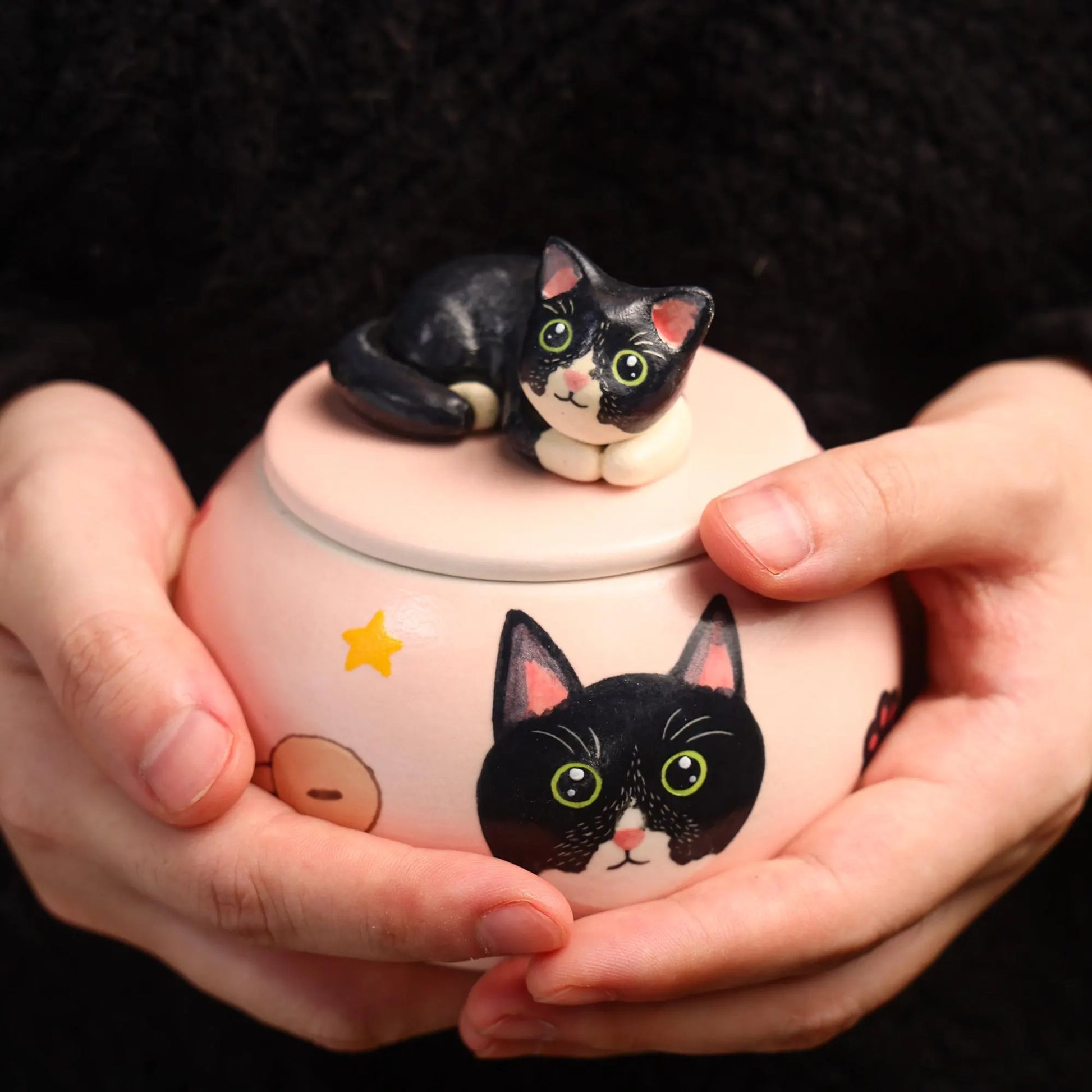 Ceramic jar with cat design held by hands against a dark background