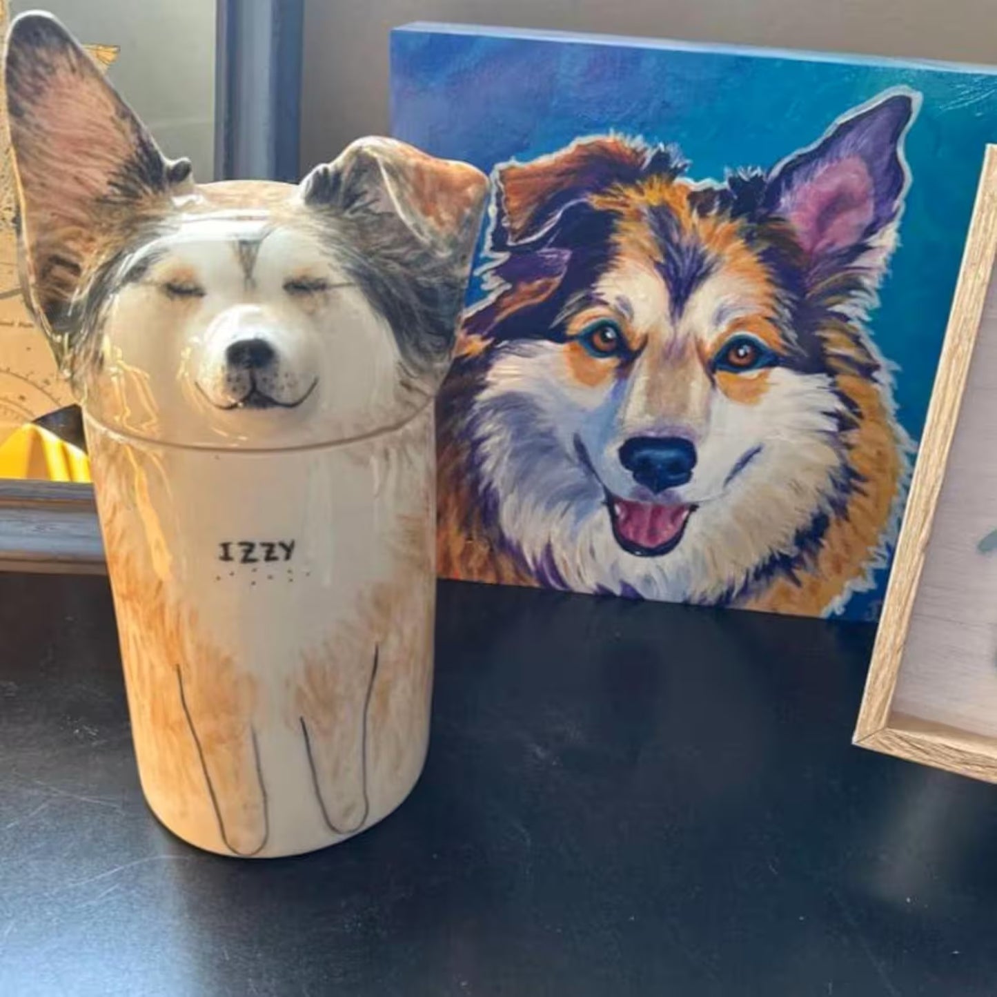 Dog-shaped TimoPaw Custom pet ceramic urn with 'IZZY' branding next to a painting of a dog on a dark surface.