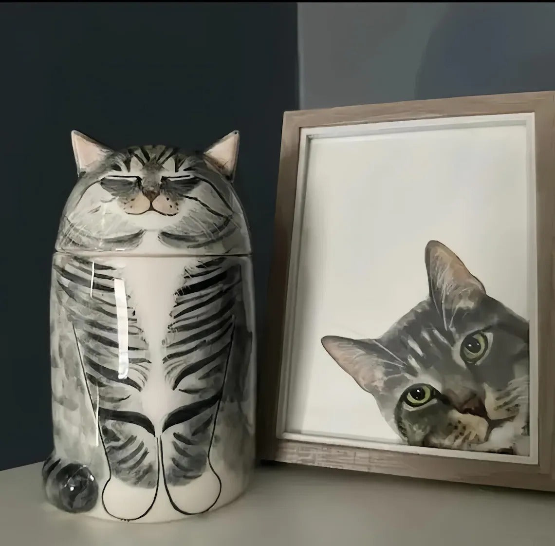 Cat-shaped urn next to a framed picture of a cat on a dark background